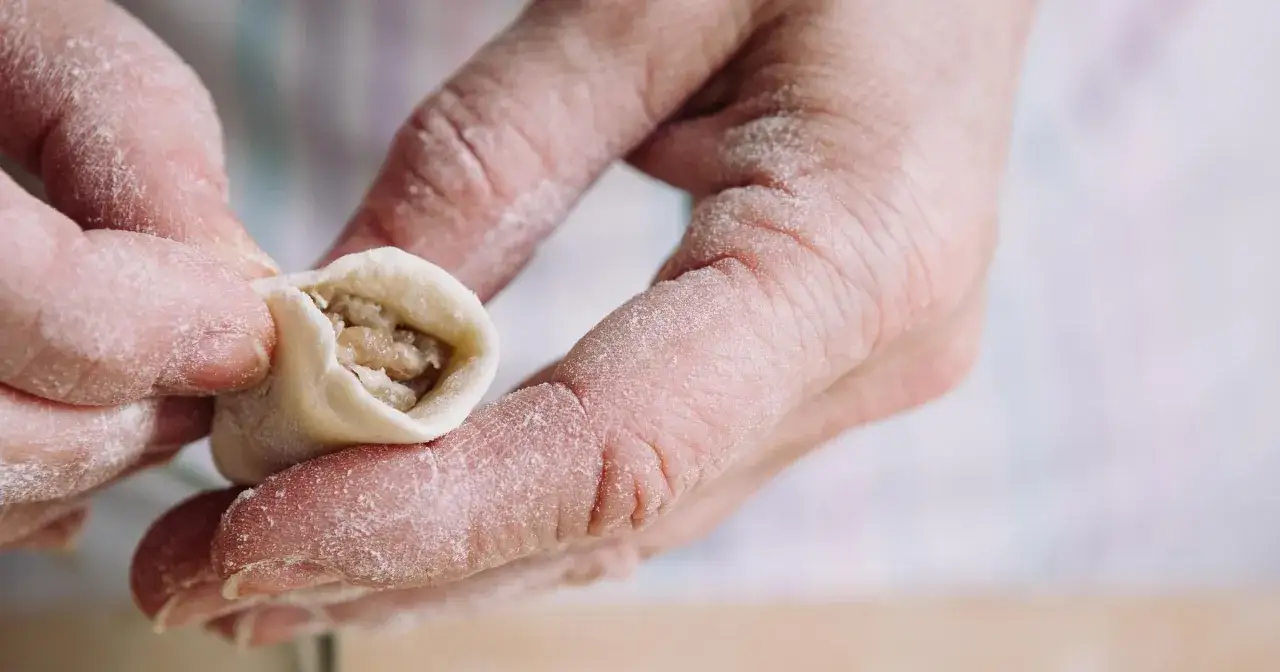 Dłonie lepią pierogi, posypane mąką. To część tradycyjnego przepisu na pierogi, który przekazywany jest z pokolenia na pokolenie.