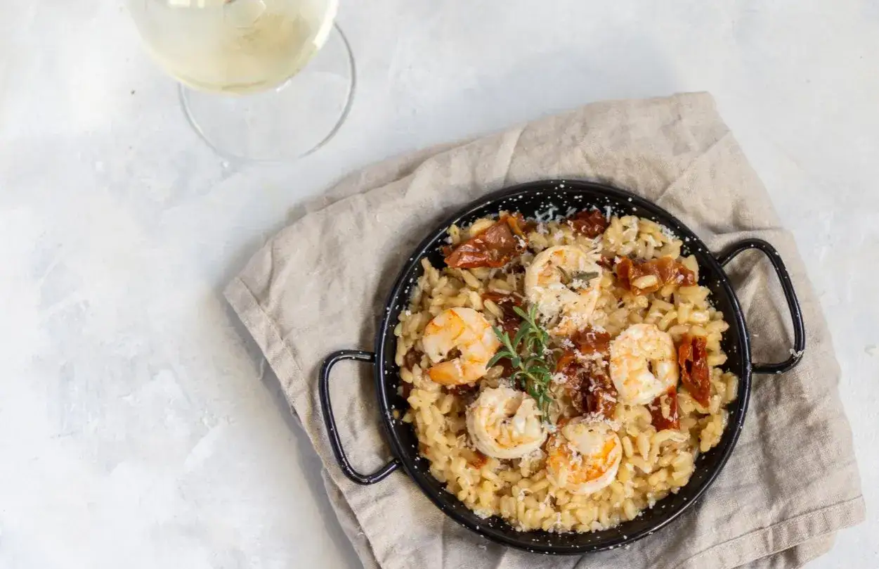 Pyszne risotto z krewetkami, suszonymi pomidorami i parmezanem, podane w czarnej patelni, obok kieliszka białego wina.
