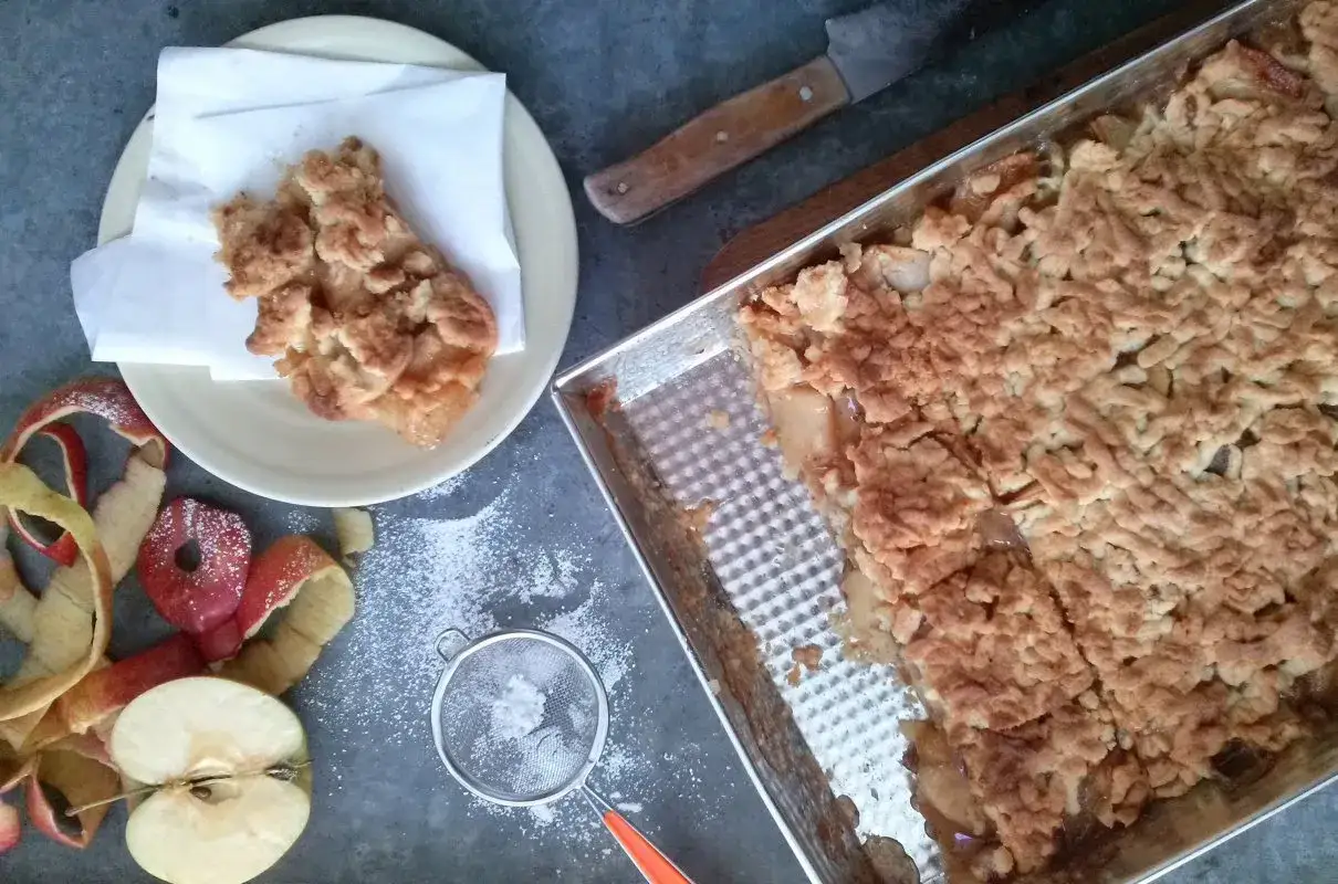 Pyszna szarlotka na dużą blachę, świeżo upieczona, z chrupiącą kruszonką. Obok jabłka i skórki, gotowe do dalszego pieczenia.