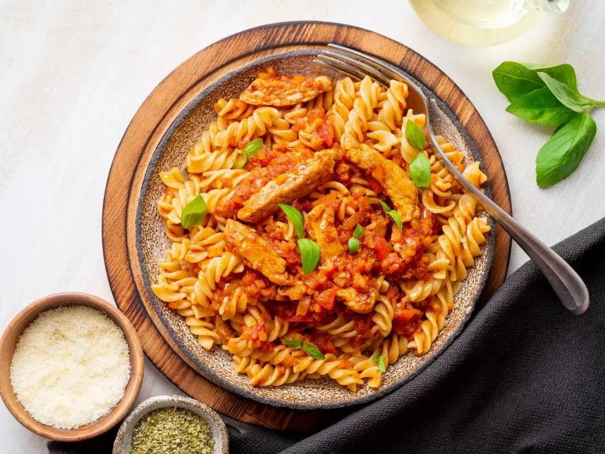 Pyszny makaron z kurczakiem i suszonymi pomidorami, udekorowany bazylią. Obok miseczki z parmezanem i ziołami.