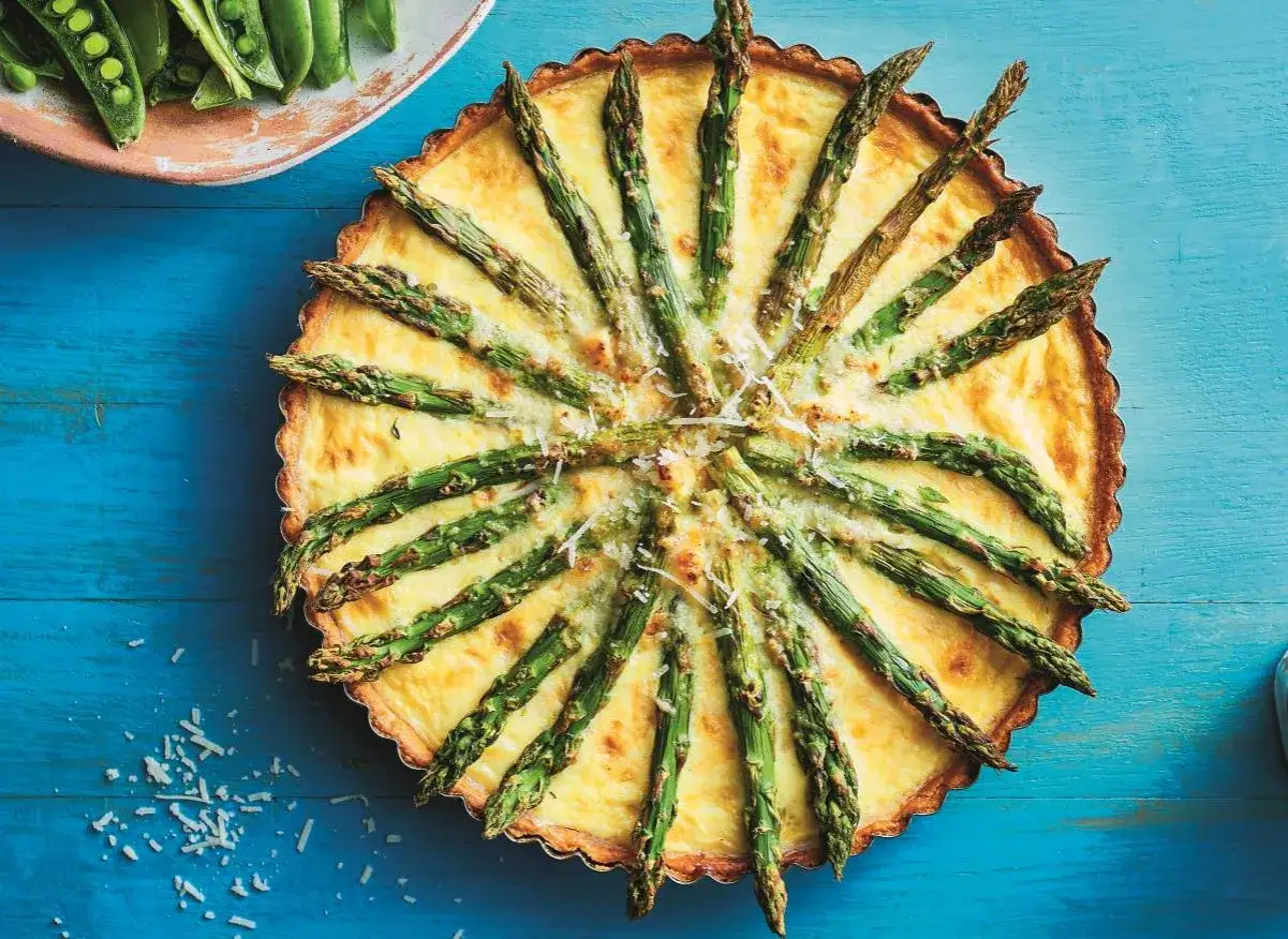 Tarta ze szparagami, udekorowana zielonymi pędami i posypana parmezanem, wygląda apetycznie na niebieskim tle.