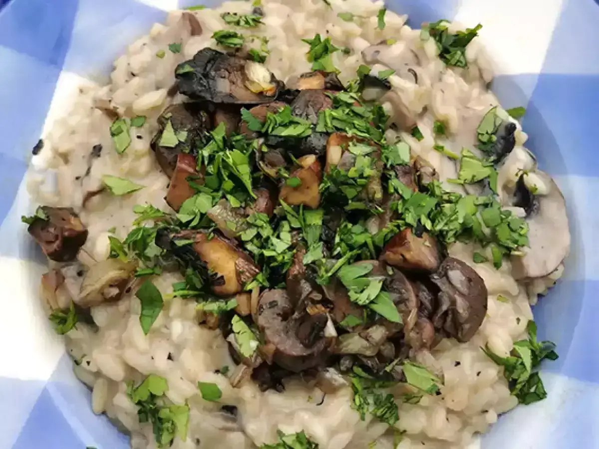 Kremowe risotto z grzybami, posypane świeżą natką pietruszki, podane w niebiesko-białej misce. Pyszne danie, kt&oacute;re zachwyci każdego miłośnika grzyb&oacute;w.