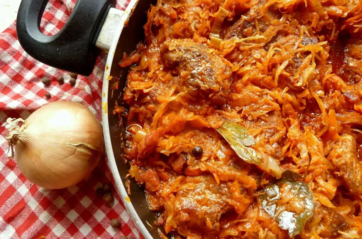 Gorąca karkówka w kapuście duszona w garnku, obok cebula i ziele angielskie na czerwono-białej kratce.