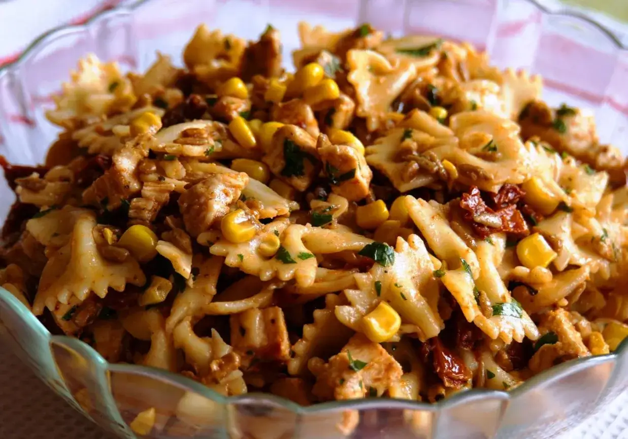 Pyszna sałatka z makaronem farfalle, kawałkami kurczaka, kukurydzą i suszonymi pomidorami, posypana natką pietruszki.
