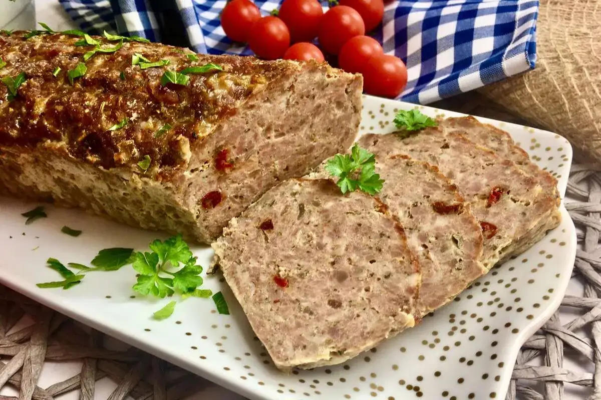 Pyszna pieczeń z jajkiem, pokrojona w plastry, ozdobiona natką pietruszki. Obok wiśniowe pomidorki i niebiesko-biała kratka.