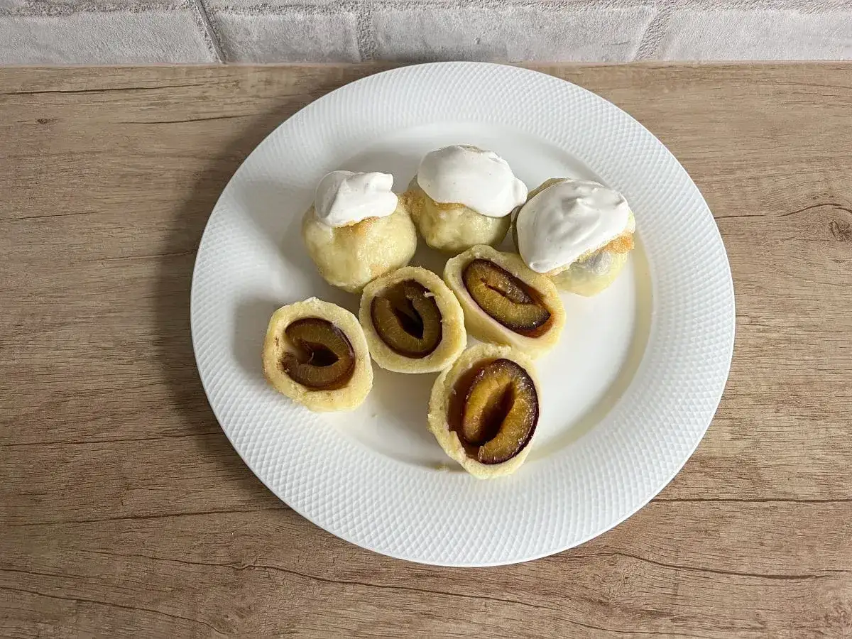 Pyszne knedle ze śliwkami, niektóre z bitą śmietaną, ułożone na białym talerzu.