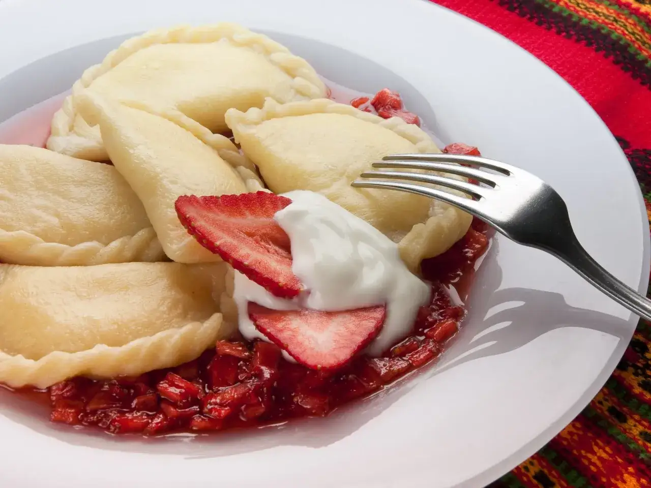 Pyszne pierogi z serem, polane sosem truskawkowym i udekorowane bitą śmietaną. Widoczny widelec czeka na pierwszy kęs.