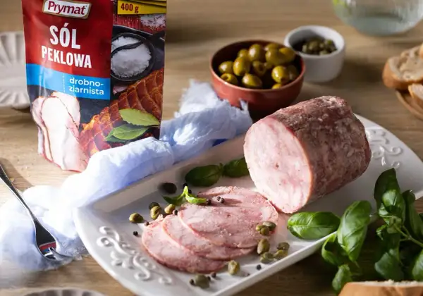 Pyszna szynka z szynkowara, pokrojona w plastry, podana z oliwkami, kaparami i świeżą bazylią. Obok opakowanie soli peklowej Prymat.