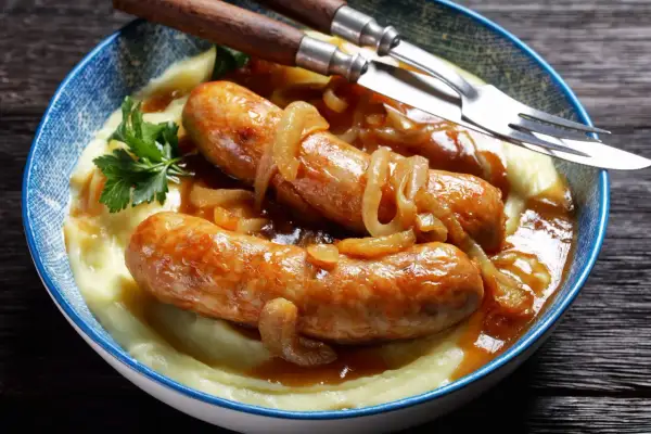 Złocista kiełbasa z cebulą i kremowym purée ziemniaczanym, udekorowana natką pietruszki. Danie podane w niebieskiej misce, gotowe do jedzenia.