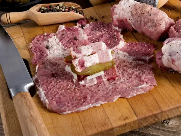 Roladki z karkówki w trakcie przygotowania. Na desce leżą rozbite plastry mięsa, doprawione pieprzem i solą, z dodatkiem boczku i ogórka kiszonego. Obok nóż i drewniana łyżka z ziarnami pieprzu.